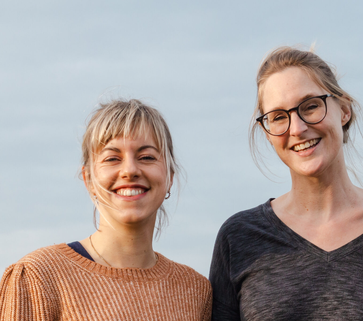 Een spontane buitenfoto van de twee vrouwelijke designers van ons designteam, glimlachend naast elkaar tegen een lichte, bewolkte lucht. Links staat een vrouw met blond haar en een pony in een warmbruine trui, rechts een vrouw met een bril en opgestoken haar in een donker shirt.